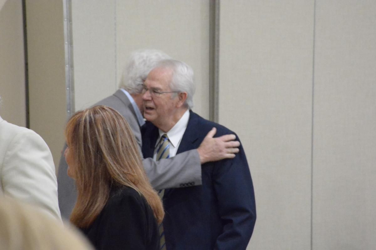 Two men in suits embracing, another person visible in the foreground. Neutral setting, possibly an event.