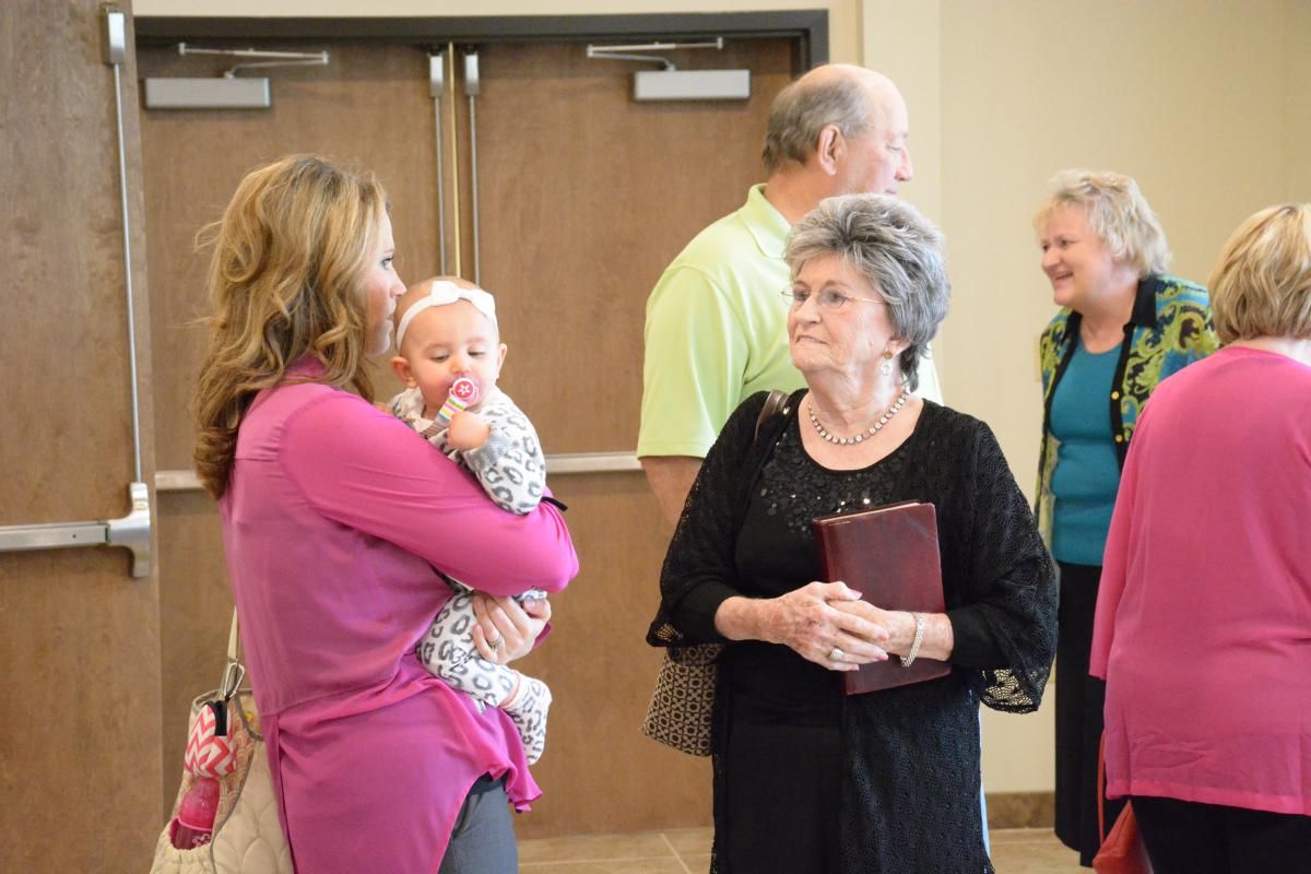 Woman holding baby talking with older woman. People in background near doors.