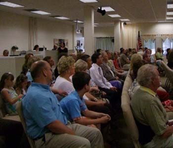 People seated in a room, likely a meeting or presentation; some looking toward the front.