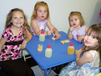 Four girls seated at a blue table with glue bottles and craft supplies, smiling.