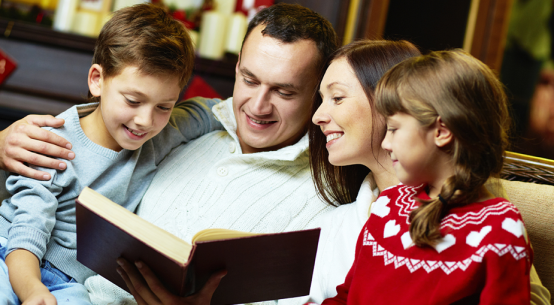 Family reading a book together; two children, parents smiling, cozy setting.