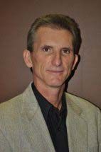 Man in light jacket and black shirt smiles, indoors.
