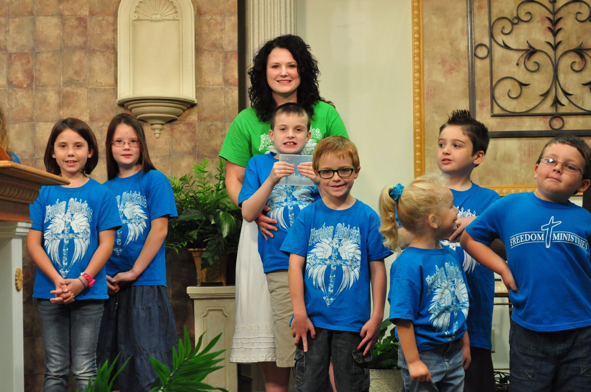 Group of children and an adult woman wearing matching blue t-shirts, posing indoors, smiling.