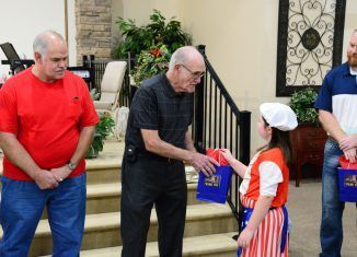 Man hands a gift bag to a girl in a patriotic outfit. Two men watch on a stairwell.
