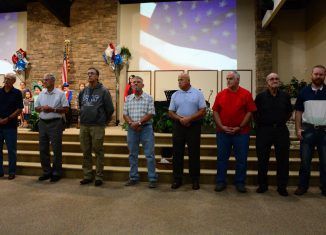 Men standing on stage in front of a US flag projection, possibly an event or ceremony.