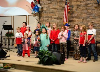 Children on stage in patriotic outfits, performing with microphone and American flag backdrop.