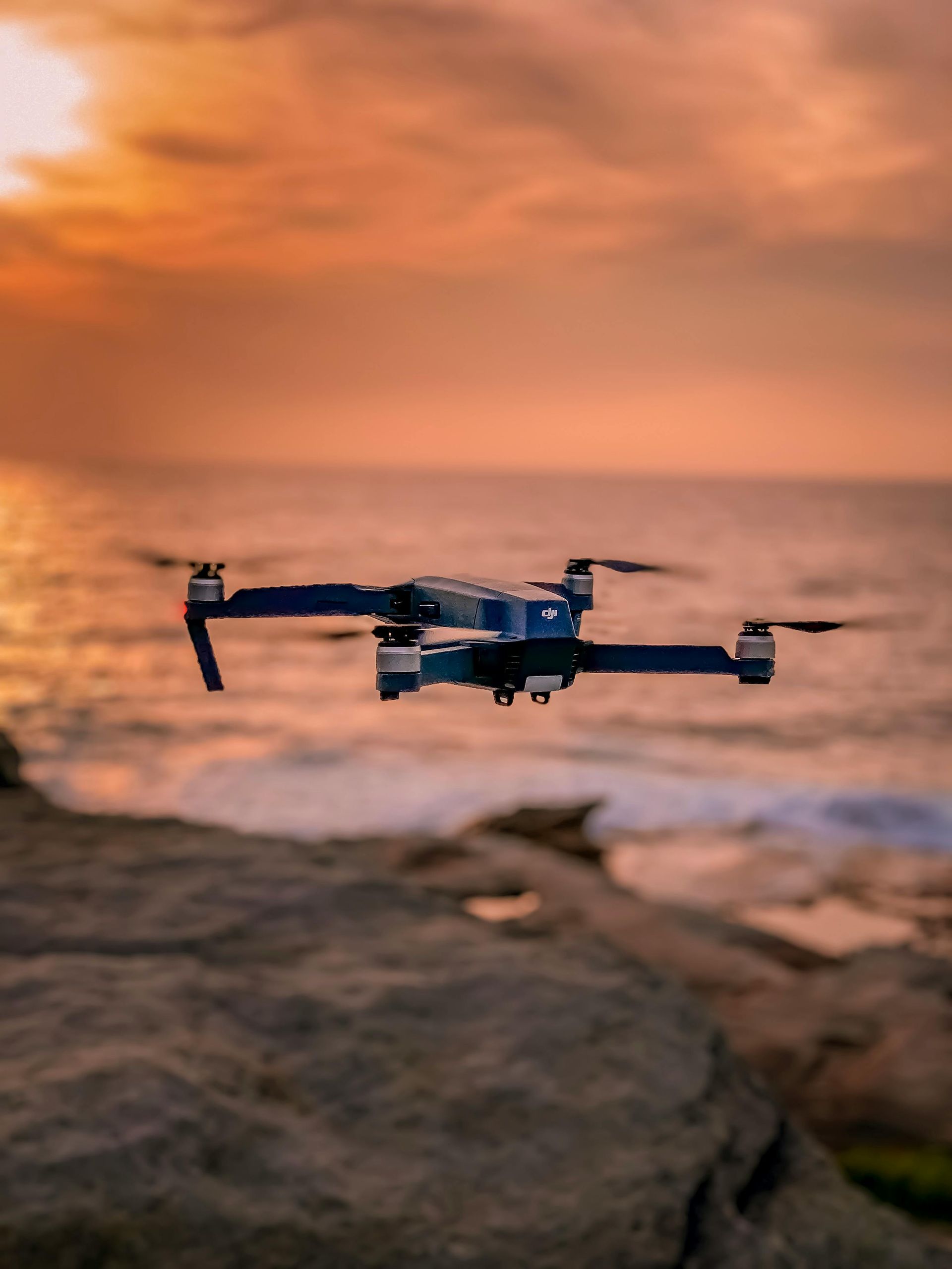 Drone vliegt bij zonsondergang boven de oceaan, met een rotsachtige kust en een kleurrijke lucht.