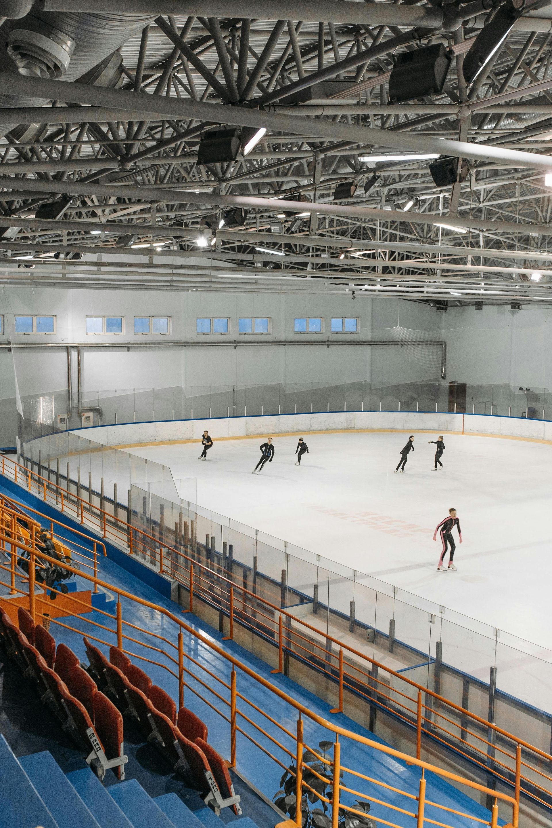 Schaatsers op een overdekte ijsbaan. Aan de zijkant bevinden zich tribunes voor toeschouwers. Plafondlampen en metalen balken zijn zichtbaar.
