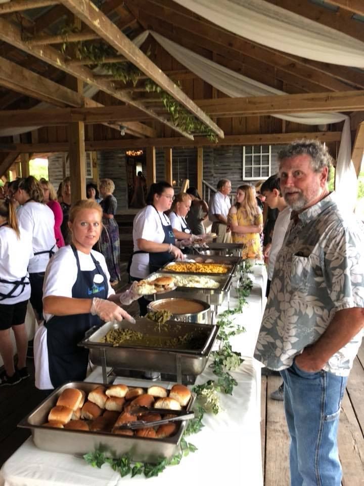 Caterers serving buffet food in a rustic hall, with a man standing beside the table in the foreground.