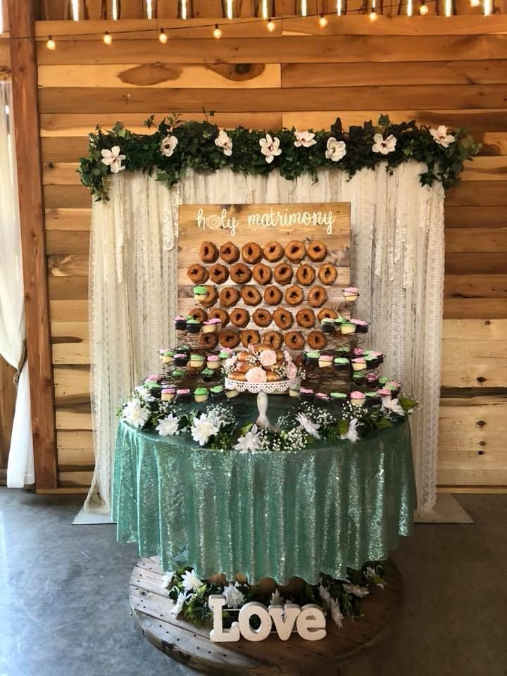 Dessert display with a donut wall, green table skirt, white drapes, and floral garland at a festive event