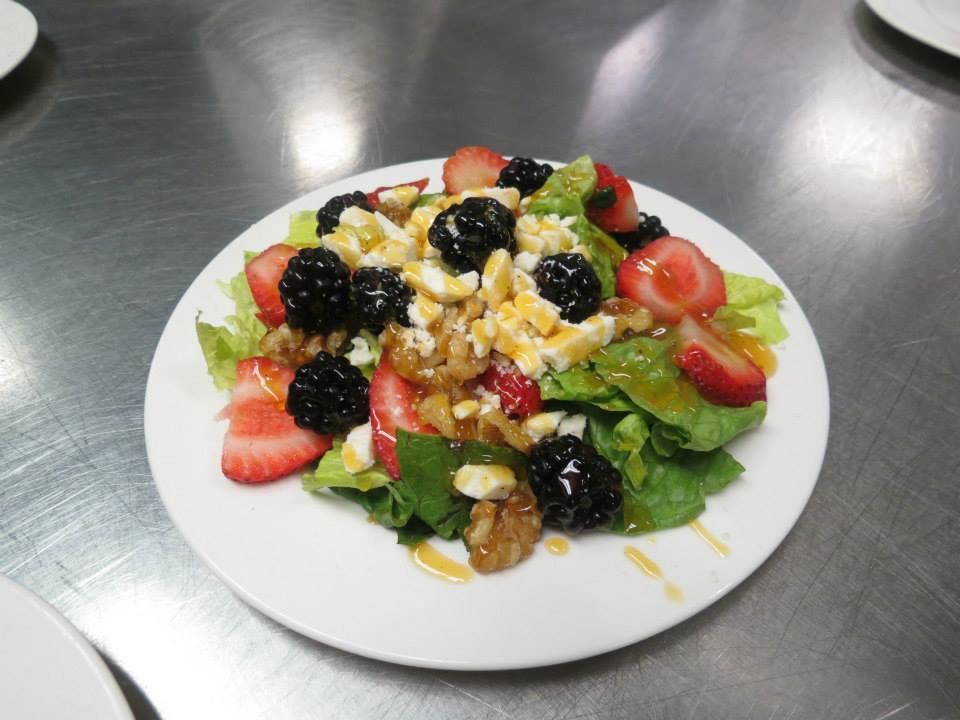 Mixed berry and greens salad on a white plate, topped with nuts and dressing on a stainless-steel table
