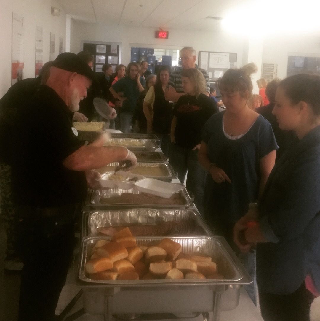 People serving food at a buffet table in a bright room with trays of bread and hot dishes