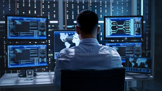 A person sits at a desk facing multiple computer monitors displaying data and maps in a dimly lit, high-tech control room.