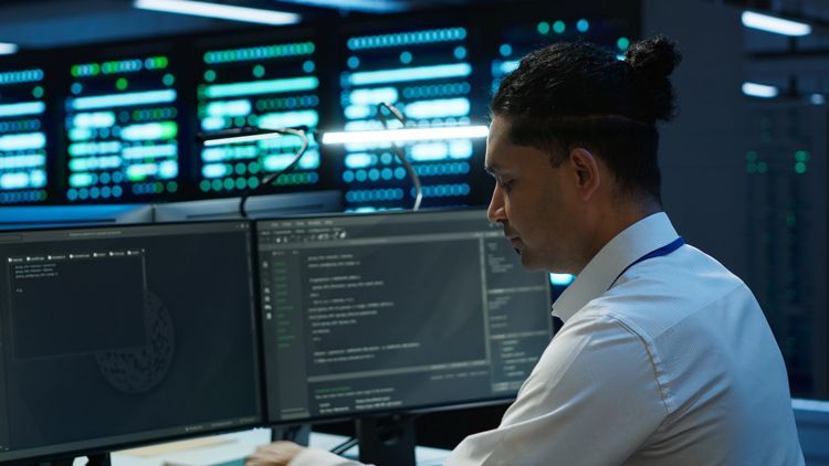 A person sits at a desk with multiple computer monitors displaying code in a dimly lit, high-tech server room.