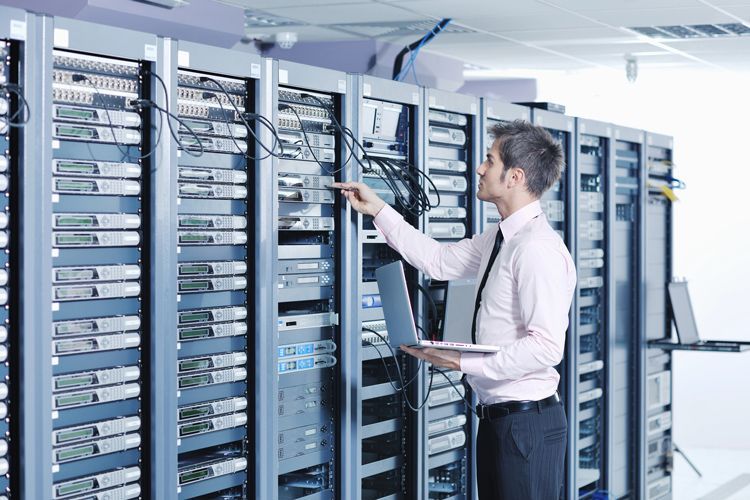 A professional in a light shirt holds a laptop while configuring a piece of equipment inside a server room rack.