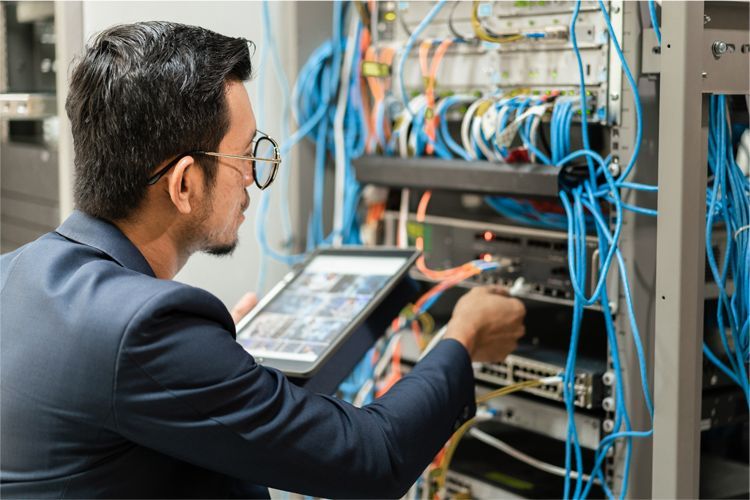 A technician in a suit examines server equipment with a tablet while managing blue cables in a network server room.