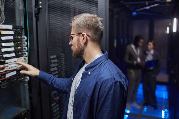A person in a blue shirt examines server equipment in a data center while colleagues converse in the background.