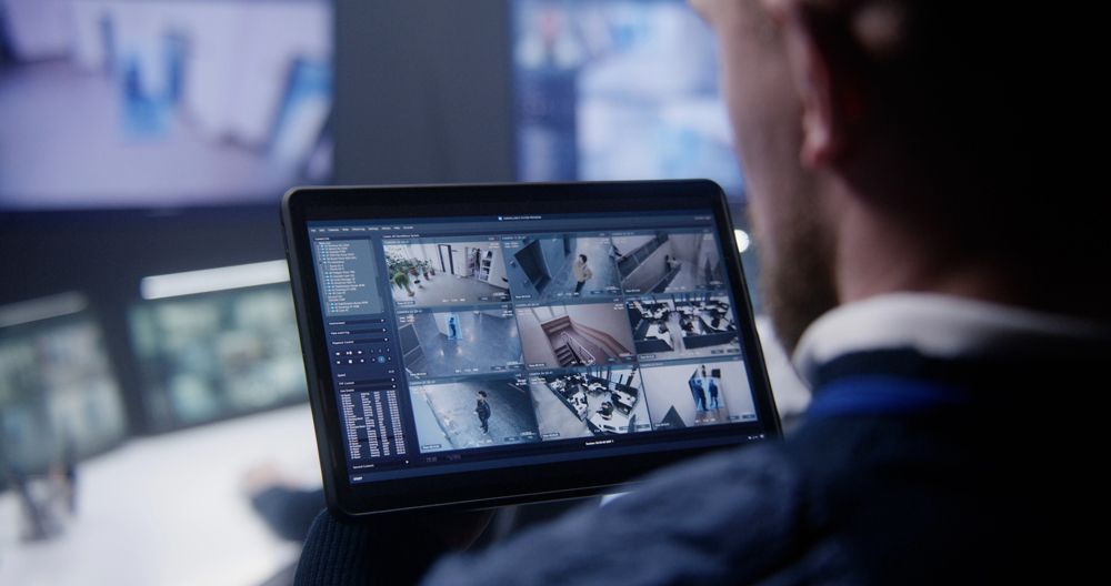 A person monitors multiple security camera feeds on a tablet in a dimly lit control room.