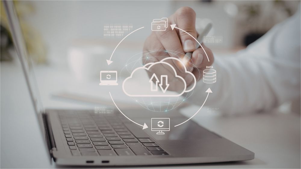 A hand uses a stylus on a laptop, overlaid with a digital graphic illustrating cloud computing and data synchronization.