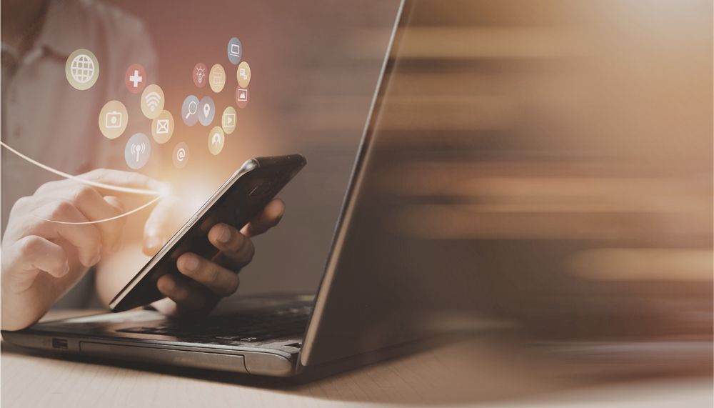A person using a smartphone near a laptop, with digital social media icons floating above the screen.