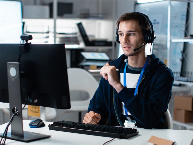 A person wearing a headset holds a stylus while sitting at a desk with a computer monitor, keyboard, and mouse.
