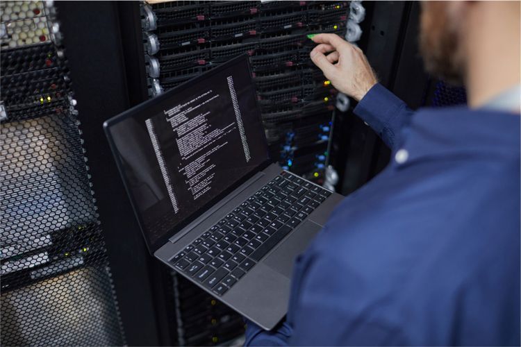 A technician in a blue shirt uses a laptop to monitor and configure server hardware inside a data center.