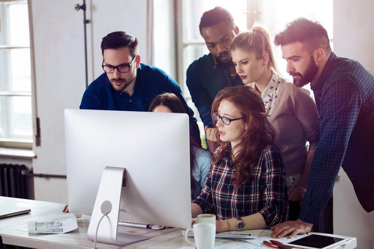 A diverse group of professionals working collaboratively around a computer screen in a bright, modern office.