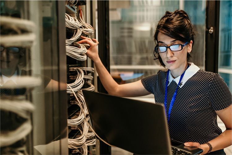 A technician wearing glasses works on a laptop while organizing cables inside a server room rack.