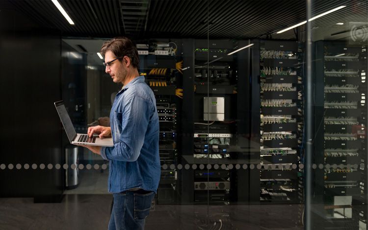 A person in a denim shirt uses a laptop while standing in a server room with glass walls and rack-mounted equipment.
