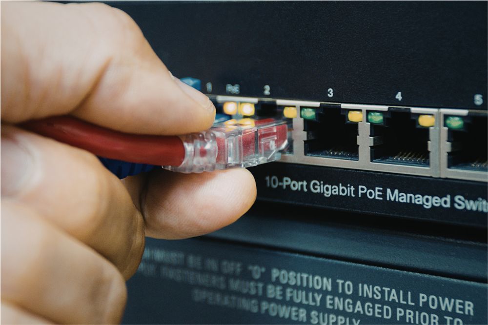 A hand inserting a red Ethernet cable into port 1 of a 10-port Gigabit PoE managed network switch.