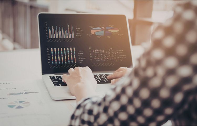 A person in a checkered shirt working on a laptop displaying various business charts and graphs at a desk.