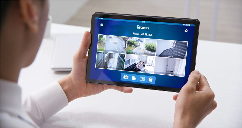 A person holding a tablet displaying a home security app with multiple live camera feeds on a white desk.