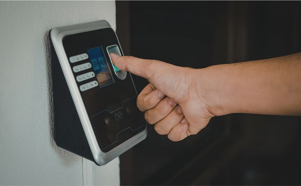 A hand pressing a finger against a biometric fingerprint scanner mounted on a white wall.