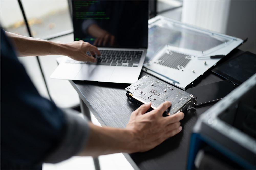 A person works at a desk, typing on a laptop with a computer component in their hand, surrounded by disassembled PC parts.