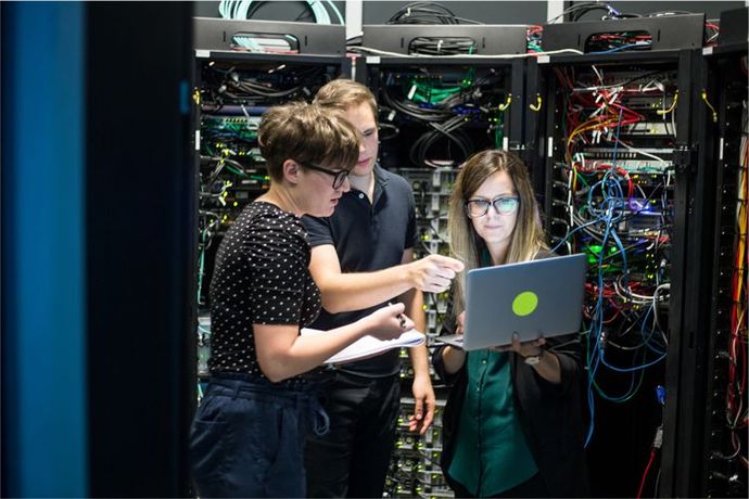 Three people stand in a server room, reviewing data on a laptop in front of racks of network cables.