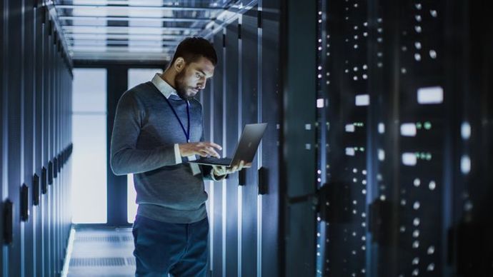 A person in professional attire uses a laptop while standing in a dimly lit, narrow server room aisle.