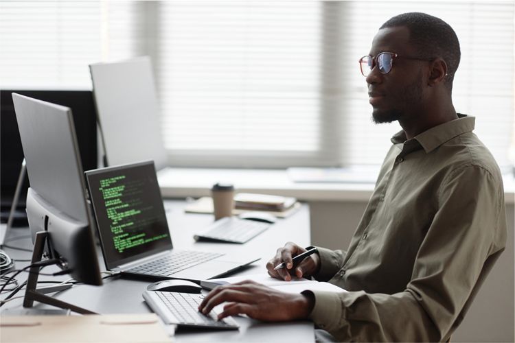 A person wearing glasses works at a desk with a laptop displaying code, a monitor, and a keyboard in a bright office.