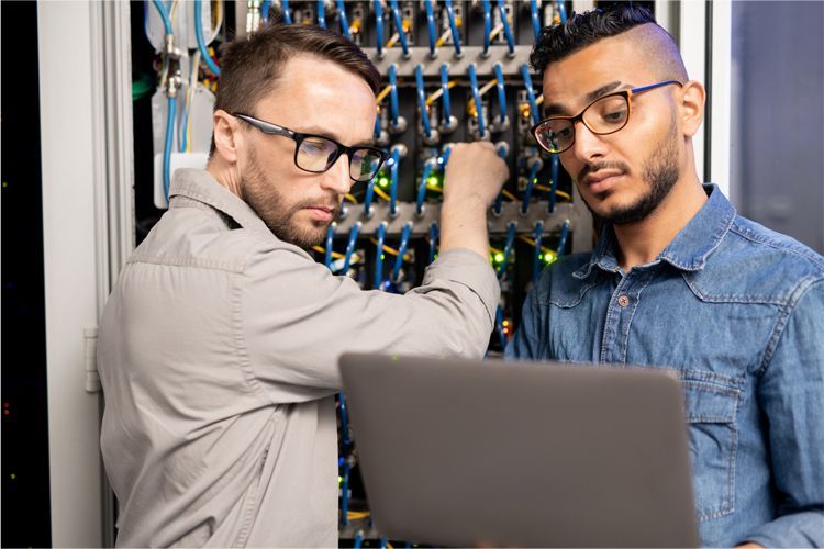 Two technicians wearing glasses work together in a server room, examining a laptop while one adjusts cables in a rack.