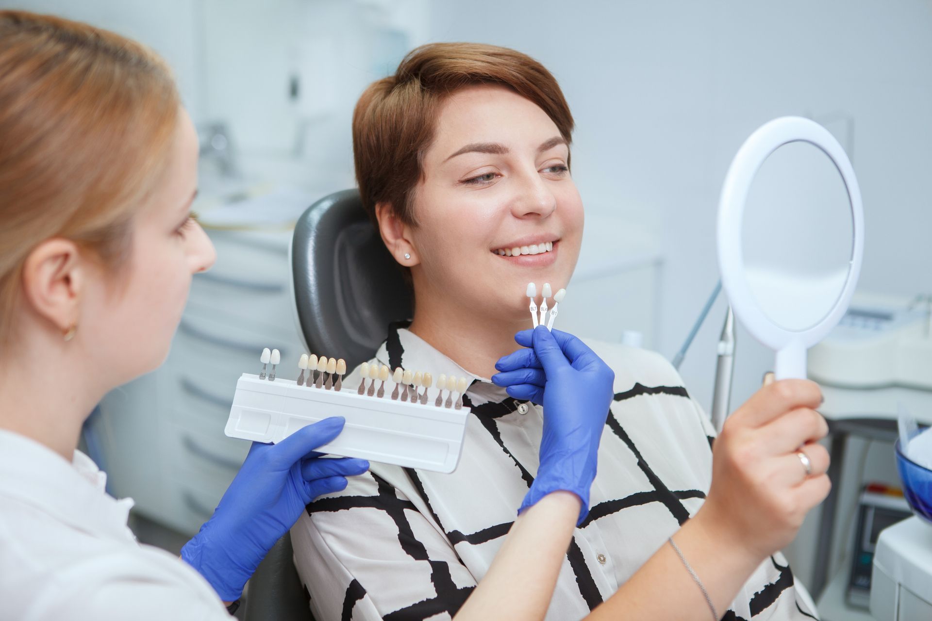 Dentist showing teeth shades to a woman patient, comparing with a mirror, in a dental clinic.