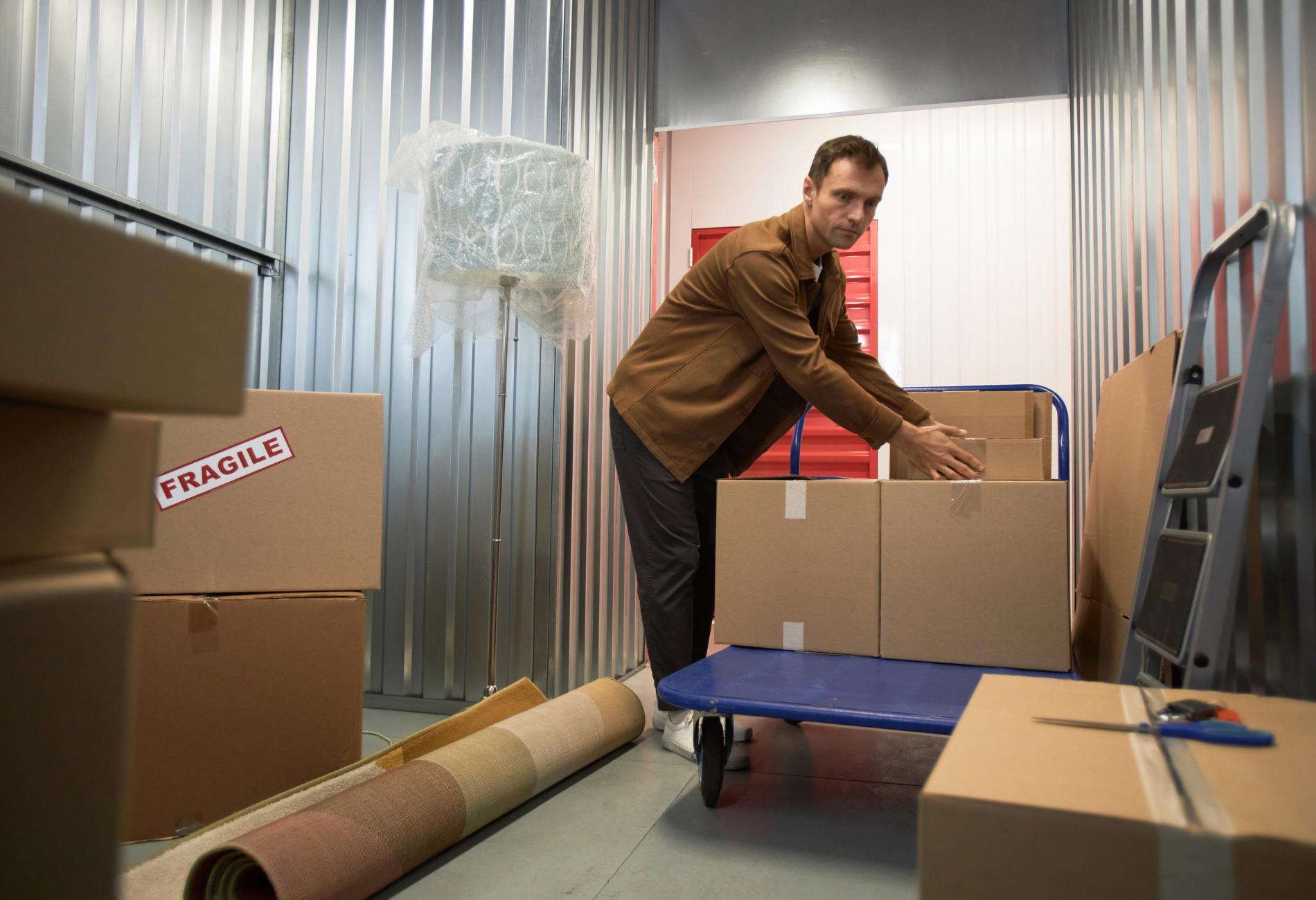 Man in storage unit packing boxes. Silver walls, tan boxes, blue cart, roll of carpet.