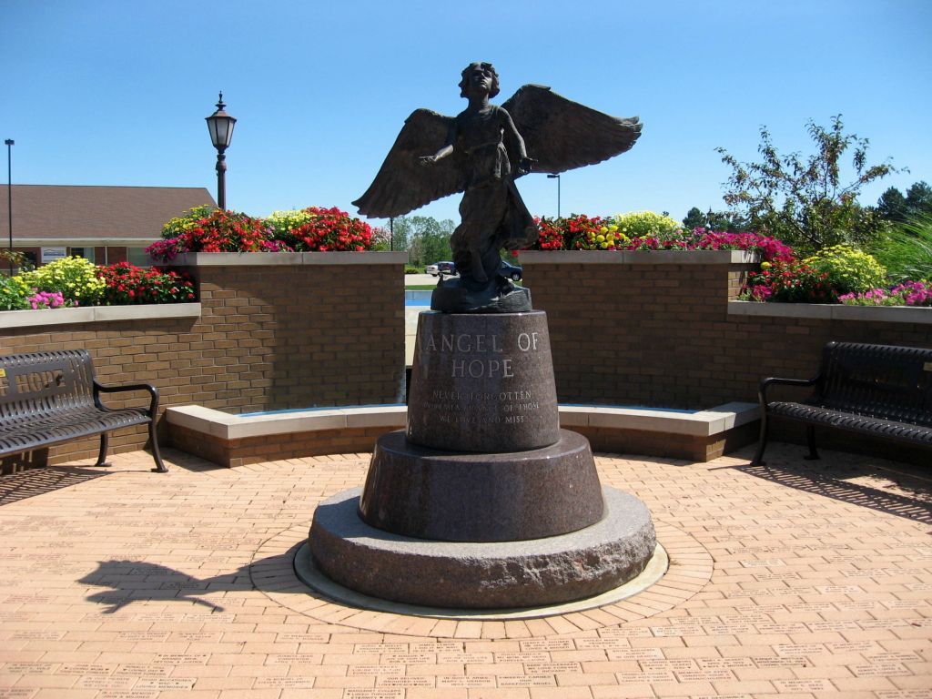 a statue of a man with wings is surrounded by benches and flowers