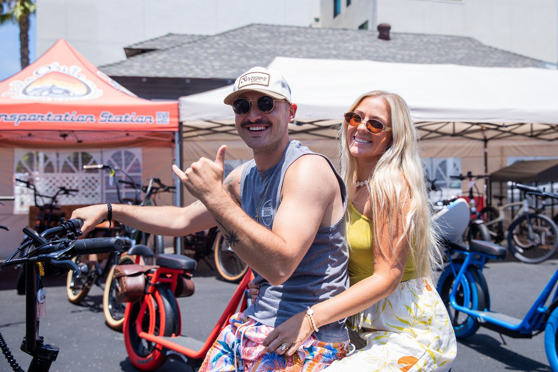 Couple on electric bikes smiling, man gives shaka. Sunny outdoor setting.