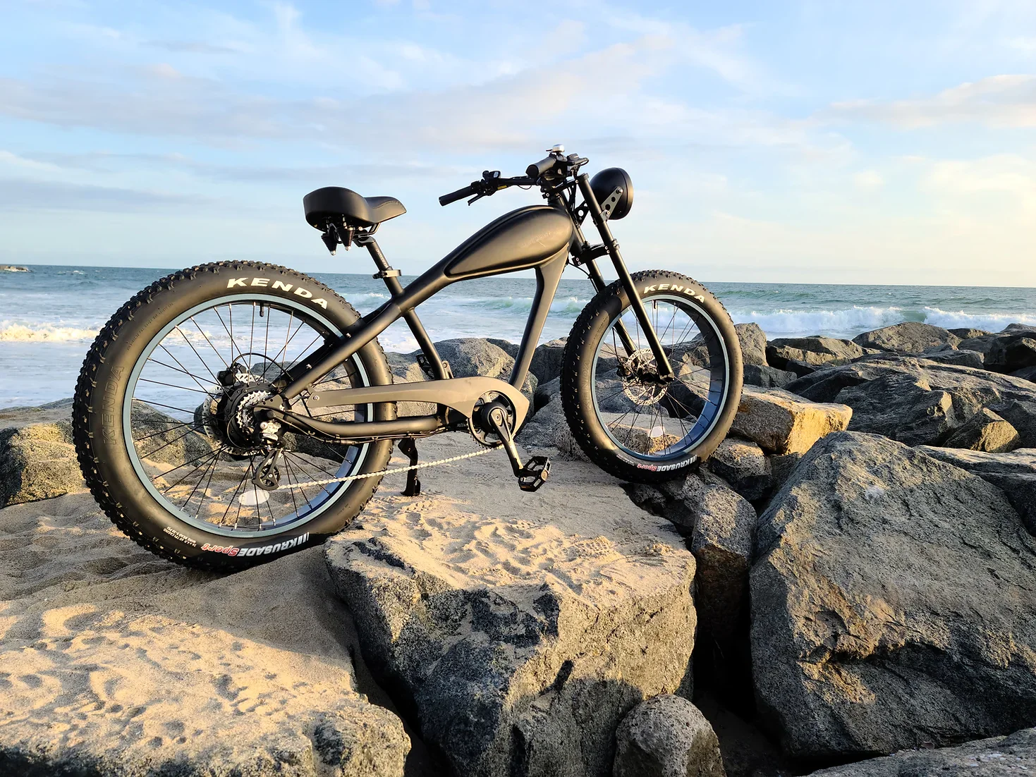 A black electric bicycle is parked on a rocky beach near the ocean.