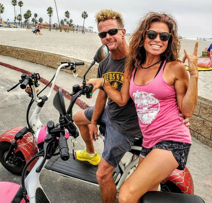 Couple on scooter by the beach, woman giving peace sign, sunny day.