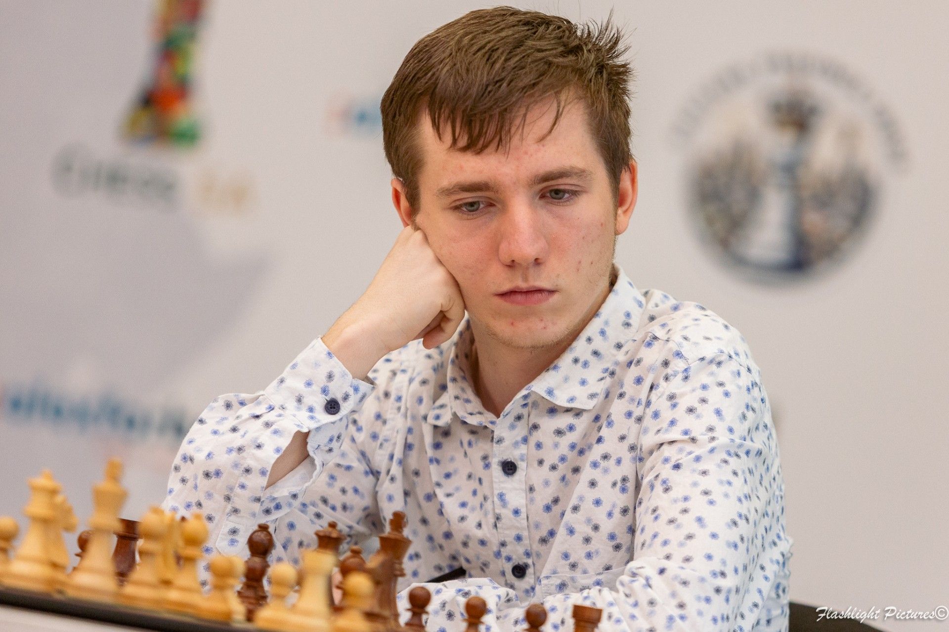 A focused chess player in a patterned white shirt sits at a chessboard with their chin resting on their hand.