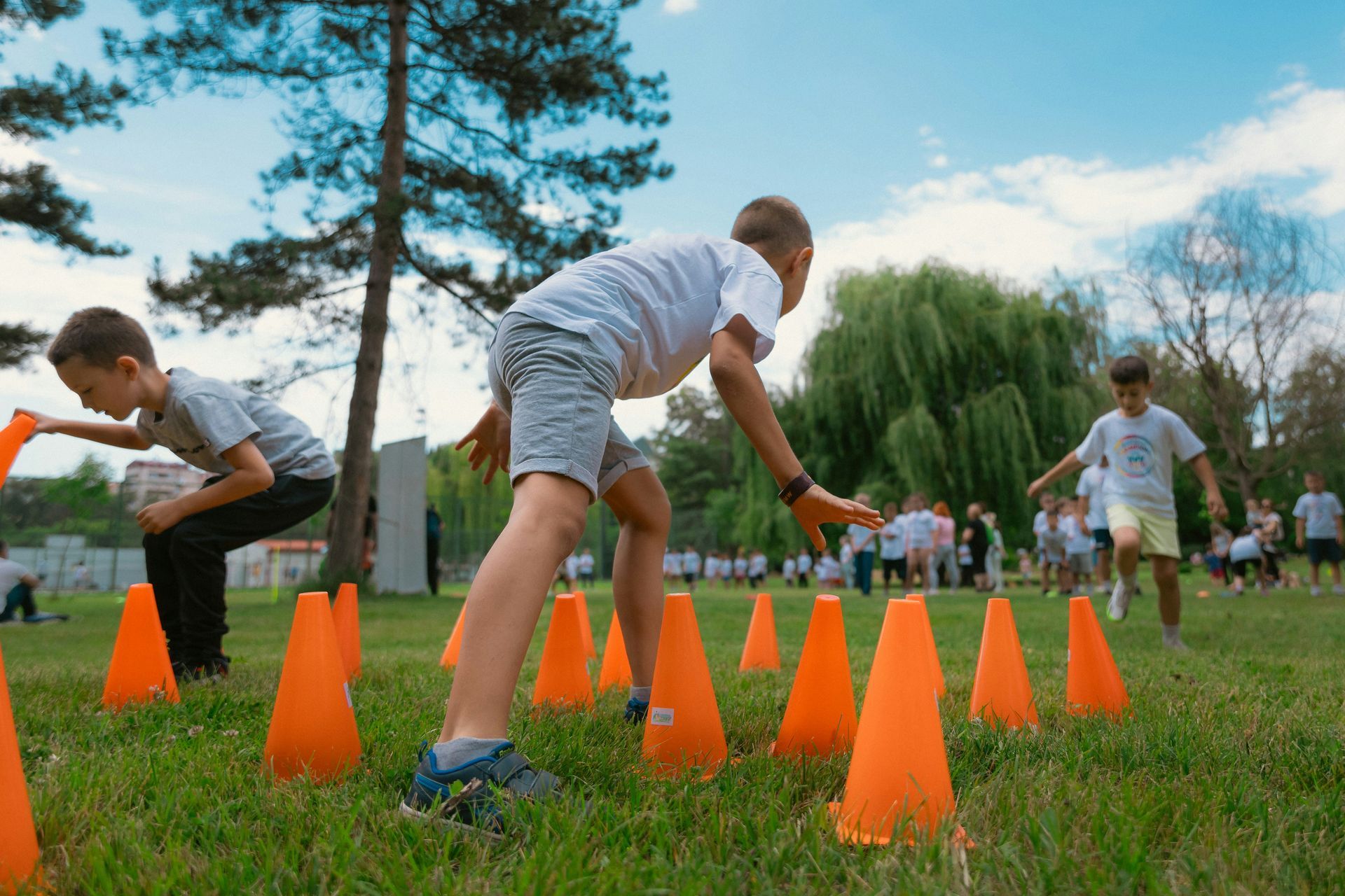 Children in white t-shirts navigate orange cones on a grassy field, participating in an outdoor activity.