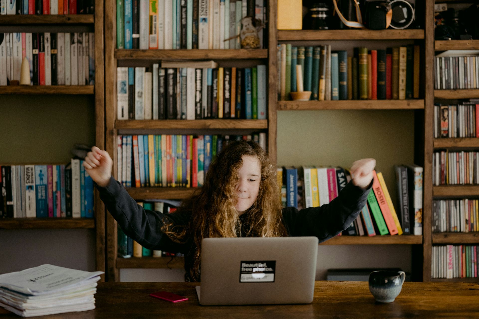 A person sits at a desk with a laptop, cheering with arms raised in front of a bookshelf filled with books.