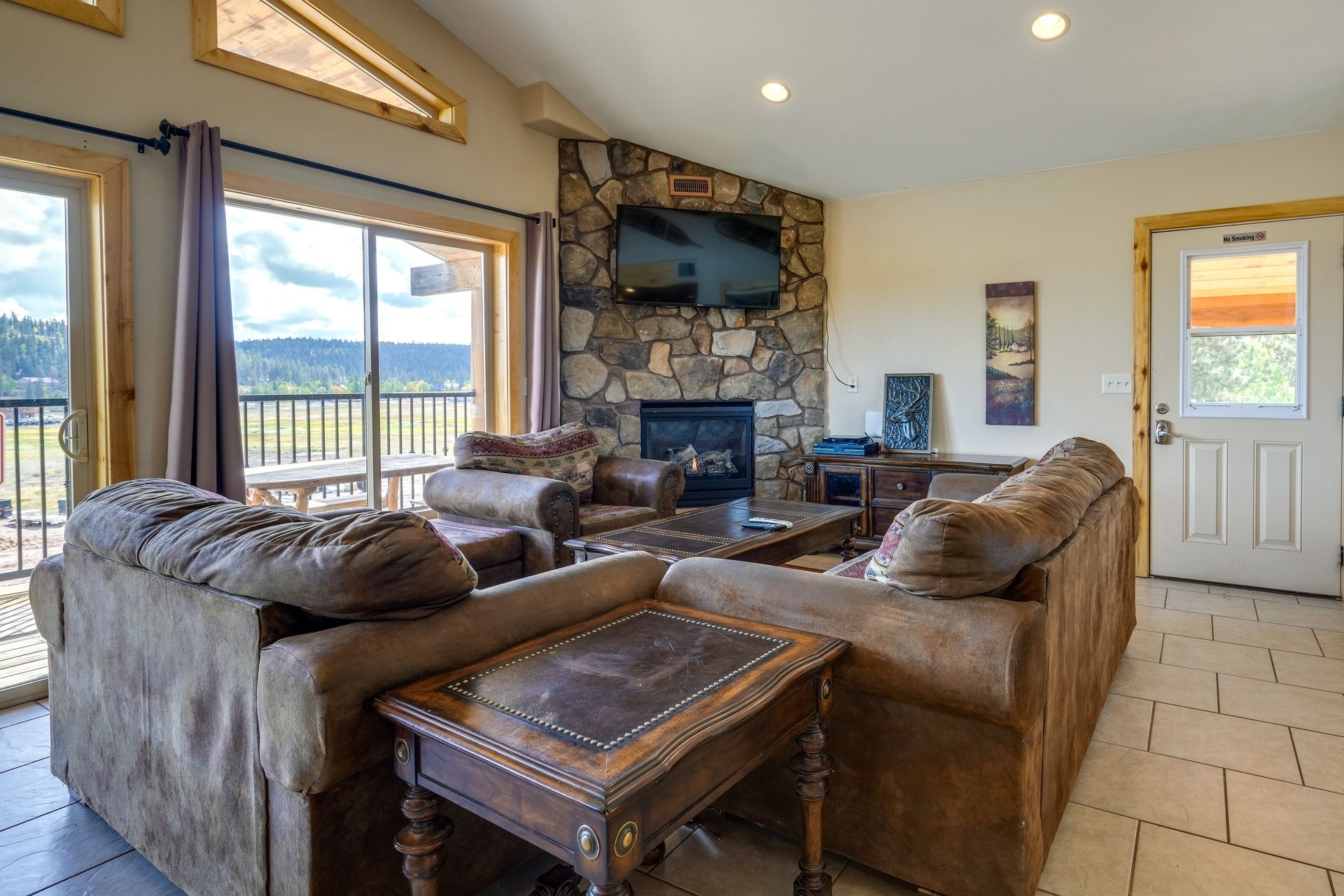 A living room with a stone fireplace, TV, and brown couches, opening to a balcony with a scenic view.
