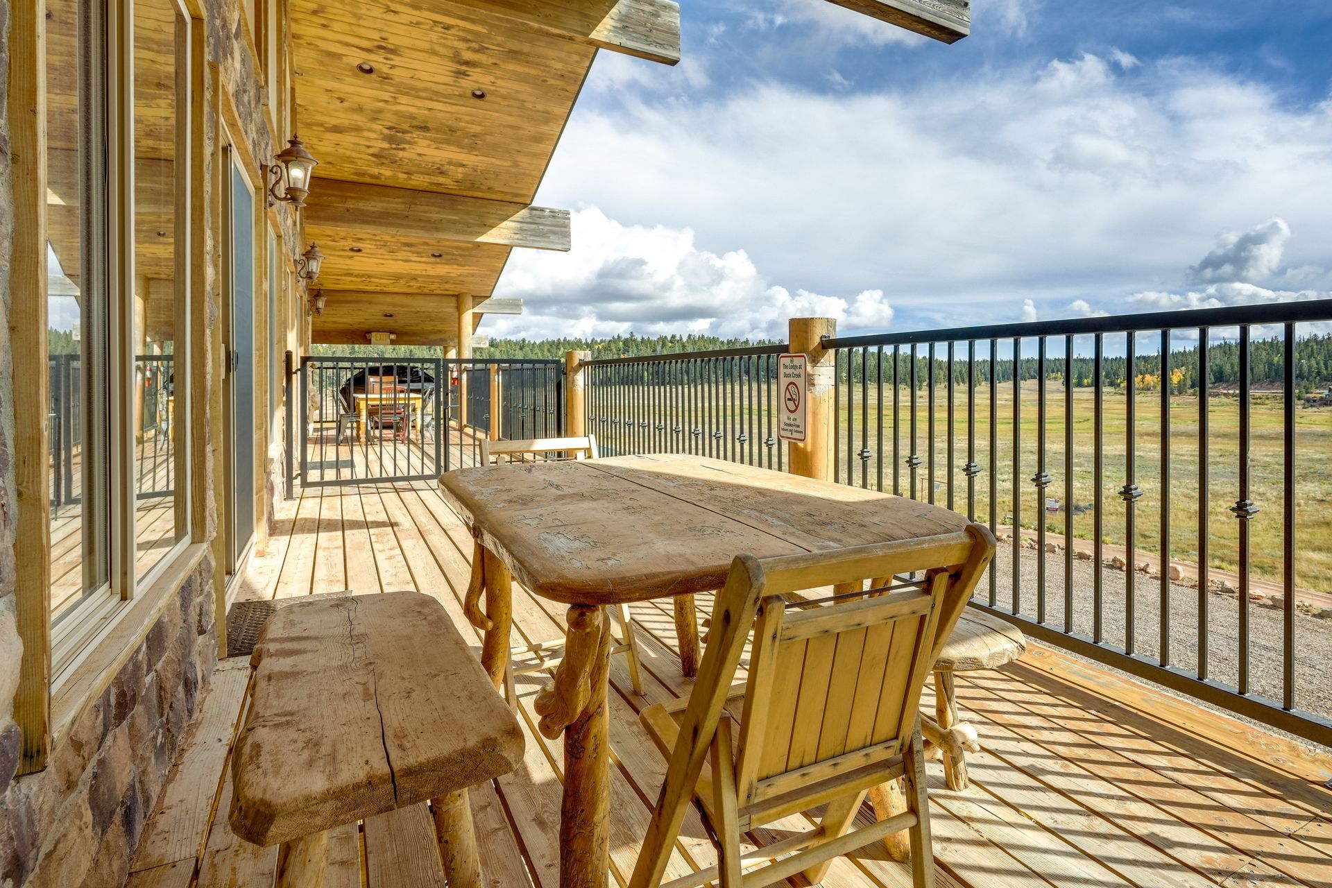 Wooden outdoor dining set on a deck with a railing, overlooking a field and trees under a cloudy blue sky.
