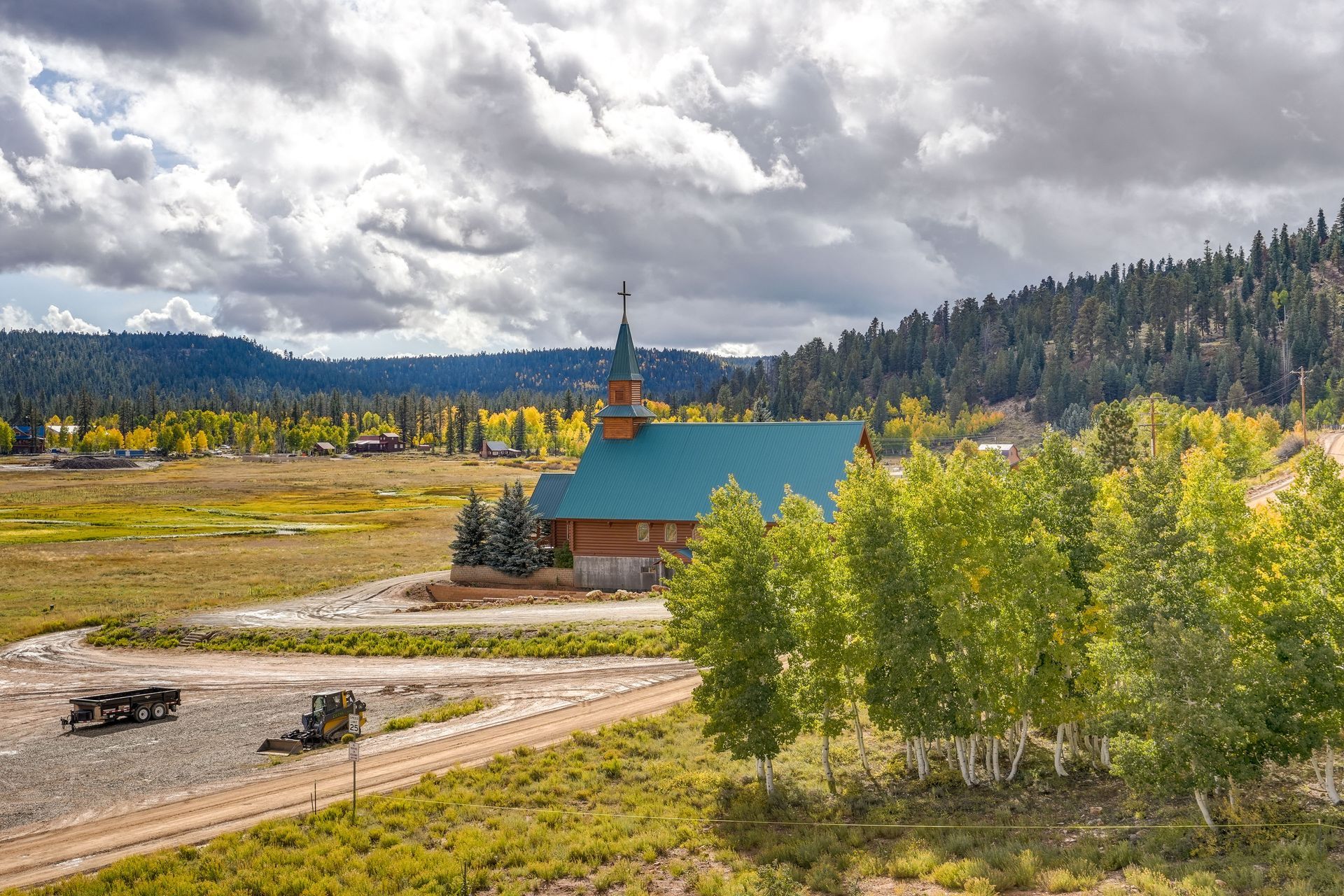 A wooden church with a green roof stands in a valley with trees and mountains under a cloudy sky.